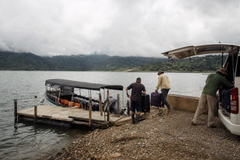 Travelers moving luggage from a shuttle to a covered passenger boat on Lake Arenal below clouded mountains