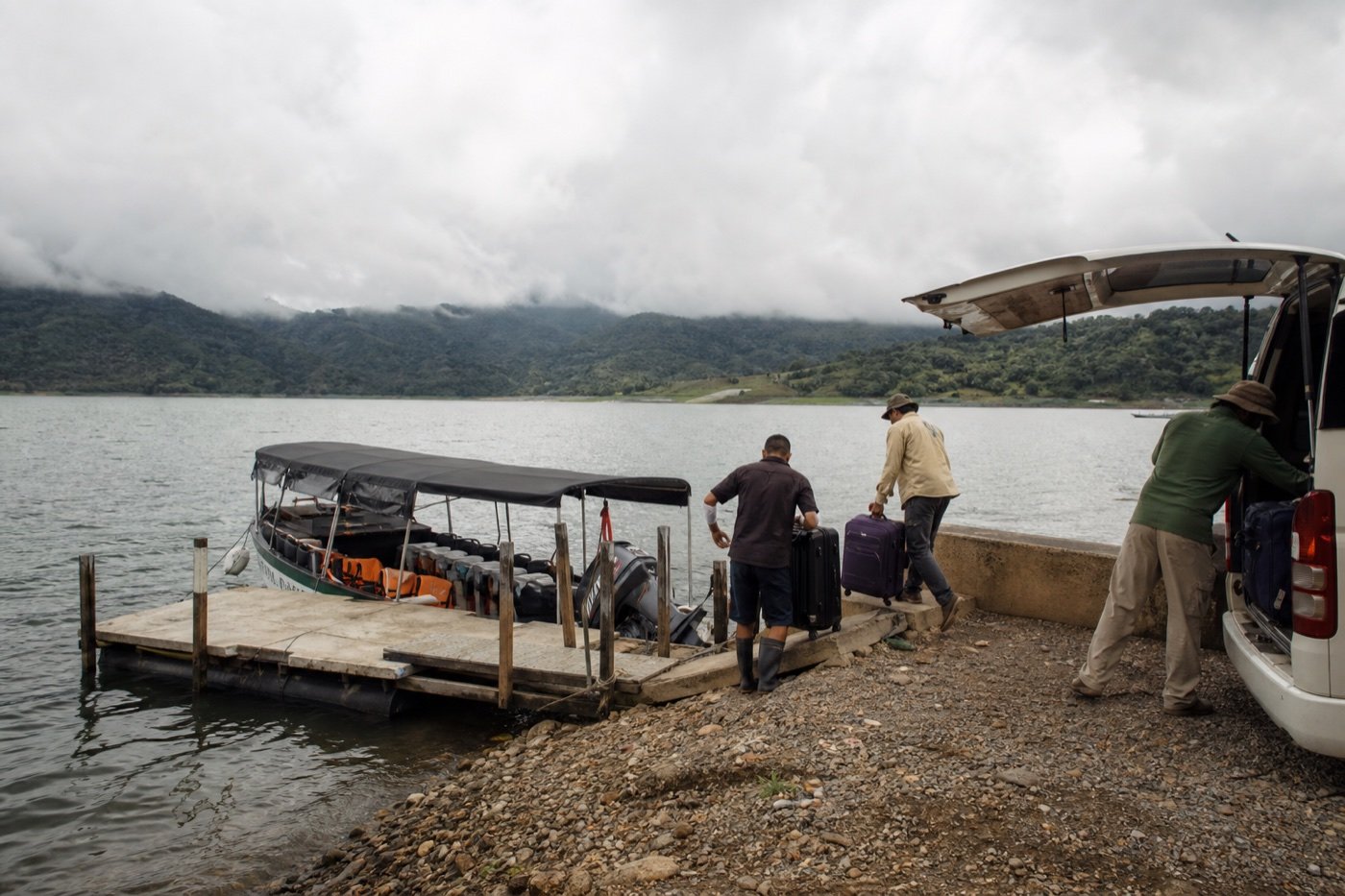 Travelers moving luggage from a shuttle to a covered passenger boat on Lake Arenal below clouded mountains