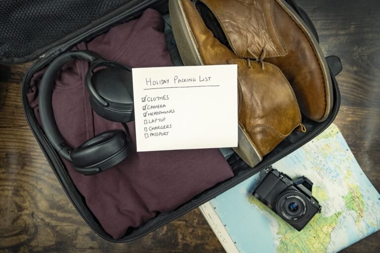 Open suitcase packed with folded clothing, shoes, headphones, and a written travel checklist beside a map and camera