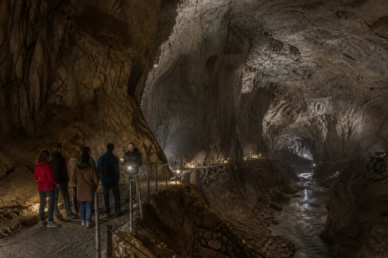 Small guided group walking through a vast illuminated Slovenian karst cavern above an underground river