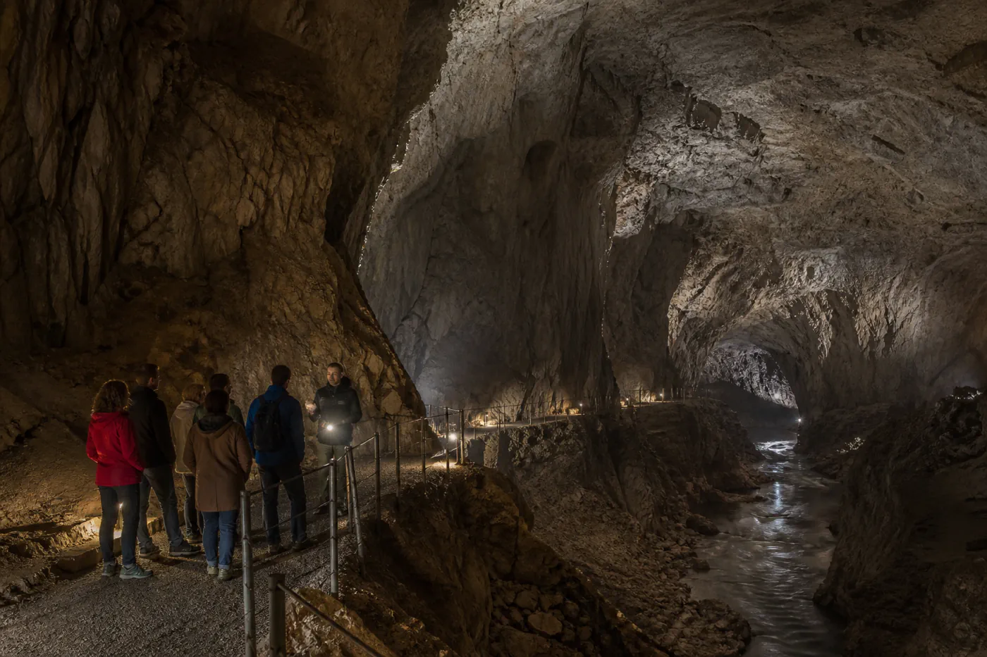 Small guided group walking through a vast illuminated Slovenian karst cavern above an underground river
